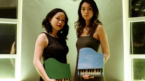 two young asian-american women in sleeveless dresses hold toy pianos as they stand under a small disco ball. 