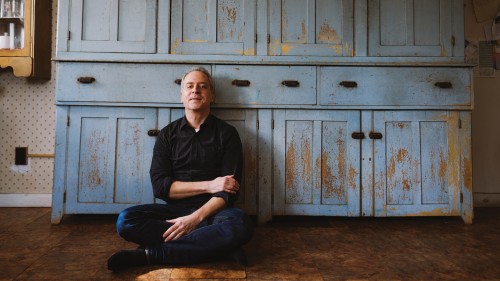 A white man in dark jeans and a black button-down shirt with the sleeves rolled up leans back against a rustic cupboard with flaking blue paint