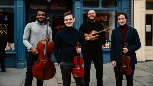 four young men smile impishly as they hold their string instruments in front of a bookstore with a bright blue facade.