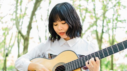 a young East Asian woman with blue-tinted wavy black hair looks down at her guitar as she plays outdoors among the trees