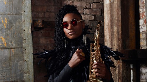 In front of a brick wall, a black woman holds a tenor saxophone. Her hair is styled with long tiny braids, she wears heavy gold-framed round sunglasses, and black-feathered epaulets on her shoulders