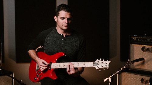 a white man looks down as he plays his red electric guitar in a music studio. His dark hair is slicked back and he has sideburns.
