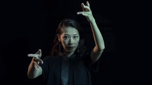 A Japanese woman, conducting, one arm raised and one gesturing forward. She looks intently ahead.