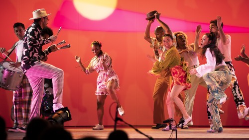 A multicultural group of five dancers clap and dance joyfully in front of a pink and orange background. 