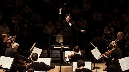 An orchestra, led by a young white man with his right arm raised and baton extended.