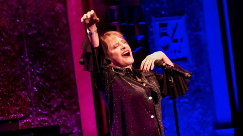 an older white woman in a sparkly top performs passionately, one arm extended and the other under her chin