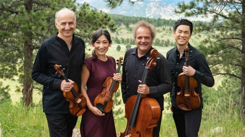 A string quartet, with two older male members who are white, and two younger members, a man and a woman of East Asian descent. They all smile warmly, in a mountain valley landscape, and hold their instruments.