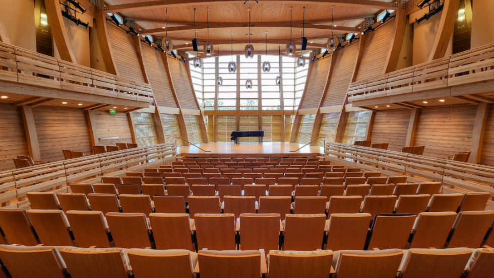 from the audience, a stage with a grand piano. All around are theatre seats, and the back wall of the stage is a panoramic window overlooking a country landscape.